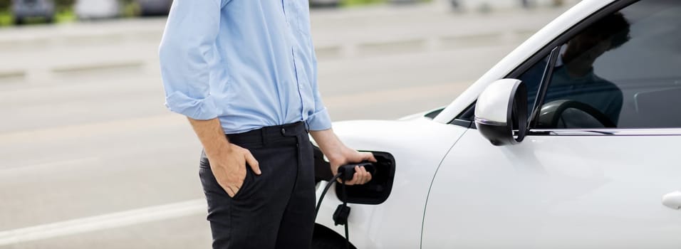 Closeup progressive man holding EV charger plug from public charging station for electric vehicle with background of residential building as concept eco-friendly sustainability energy car concept.