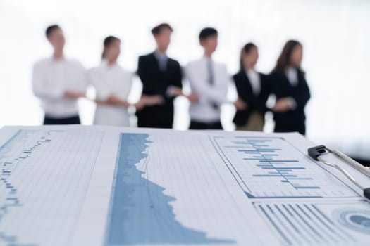 Closeup focus on financial report, performance result papers in front of camera with blur background of office workers, colleagues holding each other hands in line as concept of harmony in workplace.
