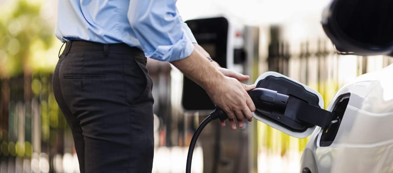 Closeup progressive man holding EV charger plug from public charging station for electric vehicle with background of residential building as concept eco-friendly sustainability energy car concept.