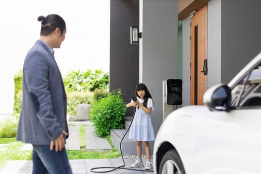 Progressive father and daughter plugs EV charger from home charging station to electric vehicle. Future eco-friendly car with EV cars powered by renewable source of clean energy.