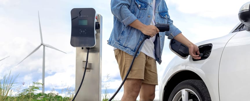 Progressive man with his electric car, EV car recharging energy from charging station on green field with wind turbine as concept of future sustainable energy. Electric vehicle with energy generator.