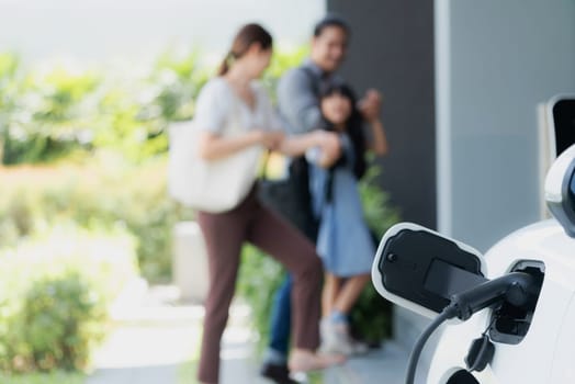 Focus closeup electric vehicle recharging battery from home electric charging station with blurred family in background. Renewable clean energy car for progressive eco awareness lifestyle concept.