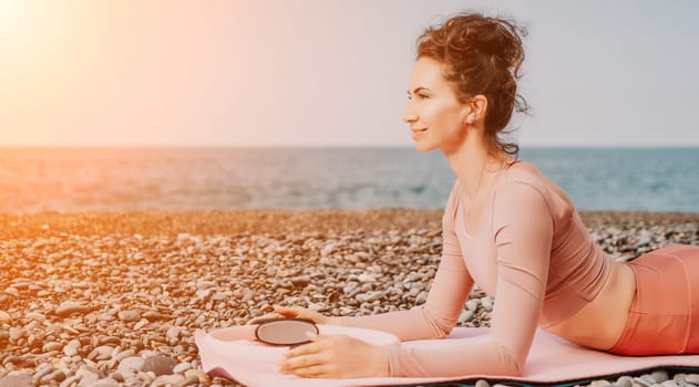 Middle aged well looking woman with black hair doing Pilates with the ring on the yoga mat near the sea on the pebble beach. Female fitness yoga concept. Healthy lifestyle, harmony and meditation.