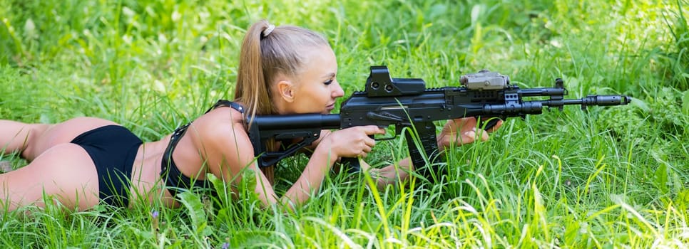 Caucasian woman in bikini lies with machine gun on green grass