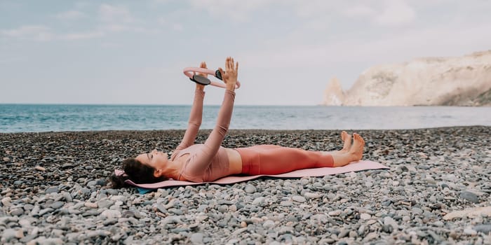 Middle aged well looking woman with black hair doing Pilates with the ring on the yoga mat near the sea on the pebble beach. Female fitness yoga concept. Healthy lifestyle, harmony and meditation.