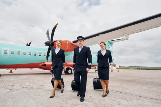 Pilot in uniform and two stewardess is outdoors near plane.