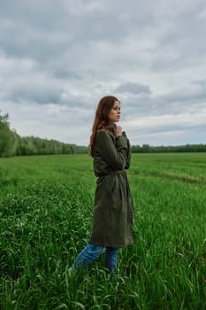 a beautiful woman in a dark coat stands in a green field in the spring in rainy weather. High quality photo