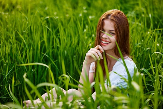 portrait of a beautiful, happy woman sitting in the grass in a light dress and smiling at the camera. High quality photo