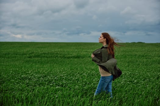 a red-haired woman stands in a green field in rainy, cold weather, holding a raincoat in the wind. High quality photo