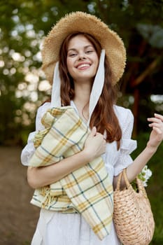 portrait of a beautiful, happily smiling woman in a light dress, a plaid in her hands and with a wicker hat enjoys outdoor recreation. High quality photo