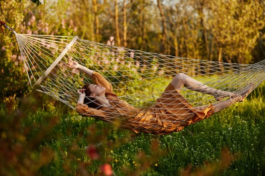 a beautiful woman is resting in nature lying in a mesh hammock in a long orange dress looking to the side, arms outstretched. Horizontal photo on the theme of recreation. High quality photo