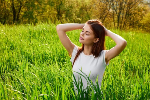 portrait of a red-haired woman in a white T-shirt walking in the grass to the waist and holding her hands near her head. High quality photo