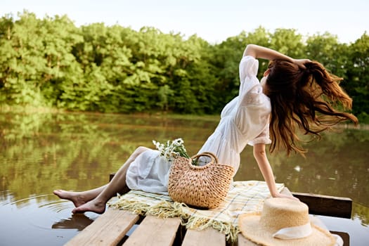 an elegant woman in a light dress sits on a pier by the lake with her legs in the water and looking at the landscape straightens her hair turning away from the camera. High quality photo