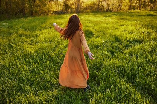 an attractive, slender, red-haired woman stands in a wide, green field during sunset in a long orange dress enjoying unity with nature and relaxation raising her arms to the sides while standing with her back to the camera. High quality photo