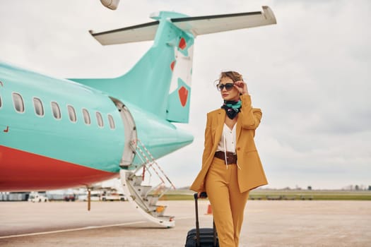 Passenger woman that is in yellow clothes, sunglasses and with luggage is outdoors near plane.