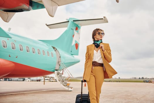 Passenger woman that is in yellow clothes, sunglasses and with luggage is outdoors near plane.