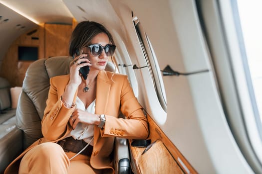 Uses phone. Young passenger woman in yellow clothes is flying in the plane.