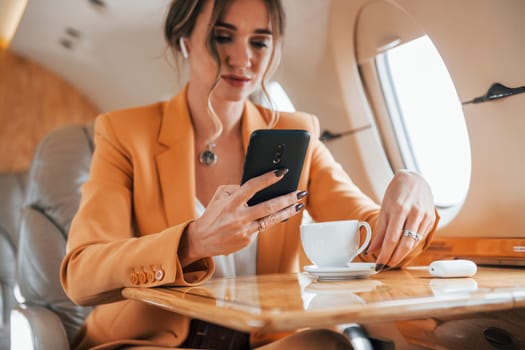 Uses phone. Young passenger woman in yellow clothes is flying in the plane.