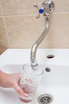 Female hand with glass. Glass is filled with transparent water under the tap with ceramic washbasin on the background