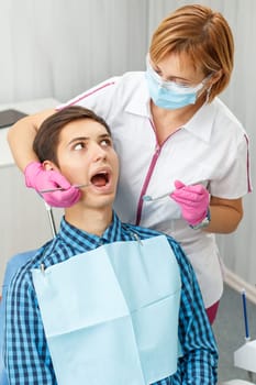 Handsome young man in visit at the dental office. He is sitting on the chair and dentist is examining patient with dental tools