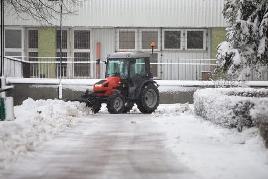 A red vehicle clears snow from the building's driveway. Removing fresh snow from the parking lot in front of the block. Winter in the city. Intense snowfall makes it difficult to drive a car.