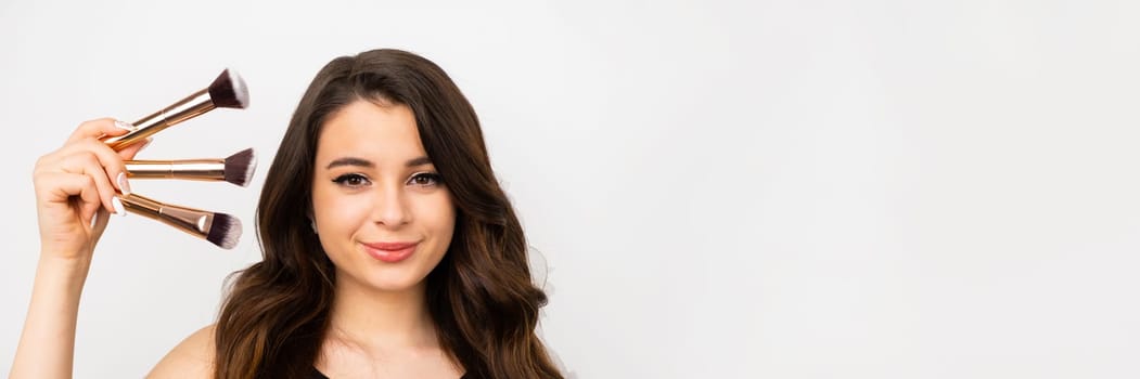 Beautiful brunette woman holding brushes near the face on the grey background with copy space.