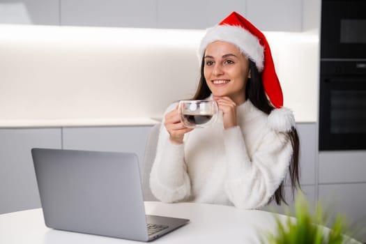 Christmas portrait of a charming brunette girl holding a cup of coffee while working at the notebook. Freelancer working on New Year and Christmas holidays.