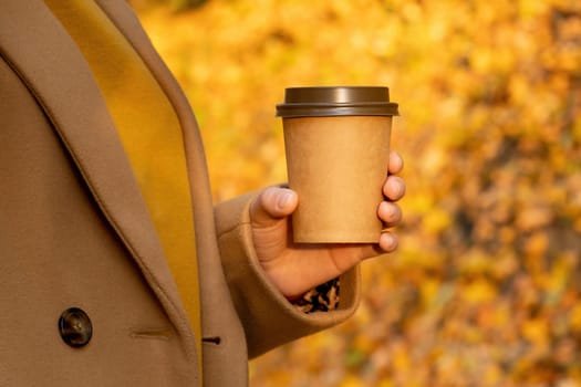 Close up woman holding a cup of coffee against a fall forest. A woman in a brown coat walks in the autumn park