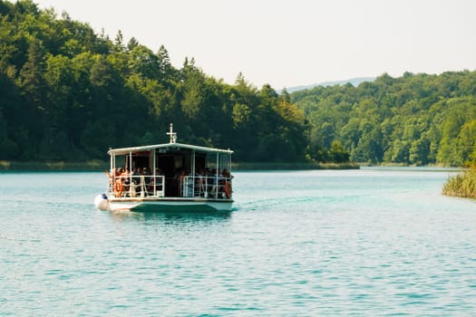 Boat with group of tourists sails on deep turquoise lake surrounded by green forestry hills. Ship excursion on blue lagoon in natural park reserve on Plitvice lakes