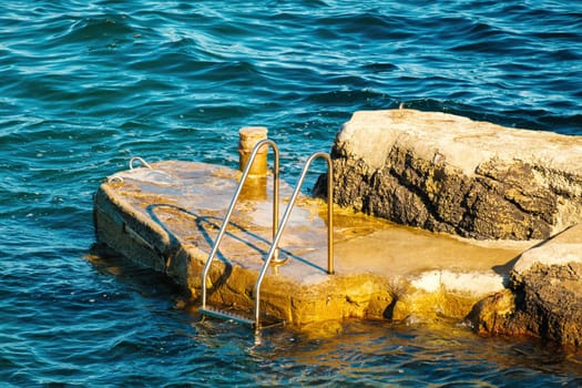 Metal ladder for swimmers descending into sea from small rock at sunlight. Stone pier washed by waves among deep turquoise sea at resort