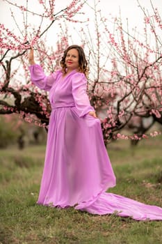 Woman peach blossom. Happy curly woman in pink dress walking in the garden of blossoming peach trees in spring.