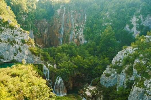 Waterfalls with cascades falling from steep rocks into canyon. Forestry and bare cliffs surround gorge with clear blue lake on bottom of Plitvice lakes upper view