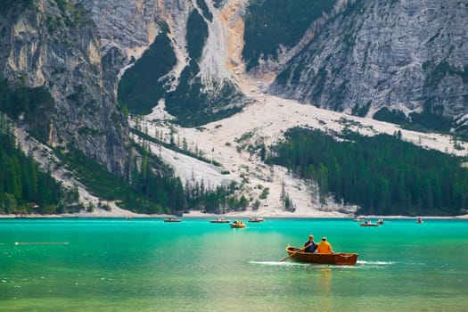 Tourists ride wooden boats on Braies lake with transparent water and Dolomites on the background in sunny day.