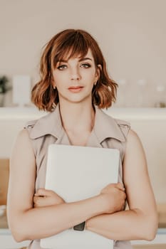 Brunette macbook. Portrait of a woman, she holds a mabuk in her hands and looks at the camera, wearing a beige dress on a beige background