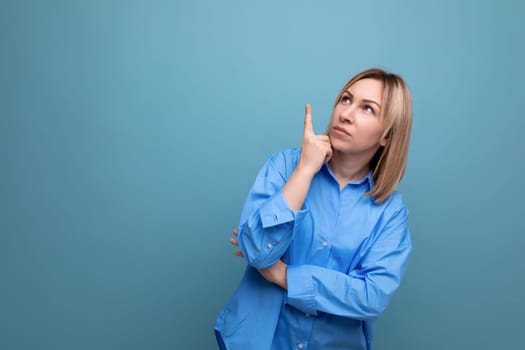 attractive young woman in casual shirt looking thoughtfully and pointing up on blue background with copy space.