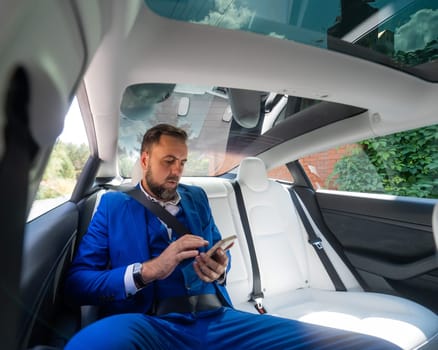 A caucasian man in a blue suit uses a smartphone in the back seat of a car. Business class passenger