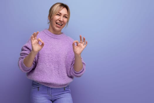 laughing blonde young woman in a lilac sweater with a kind heart on a bright background with copy space.