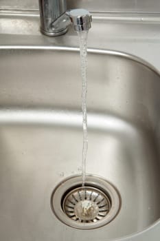 Stream of water flows from an open chrome tap with metal sink on the background. Selective focus on stream