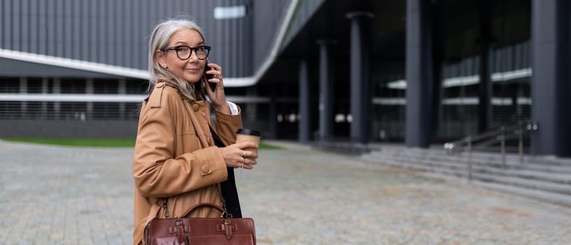 adult successful business woman walks into an office building talking on a mobile phone with a cup of coffee in her hands.