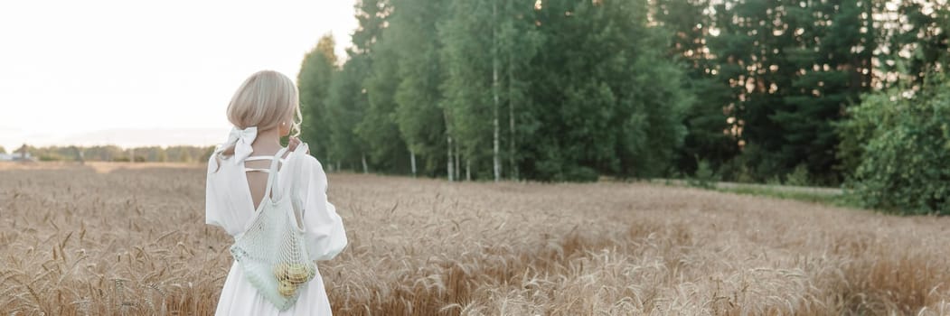 A blonde woman in a long white dress walks in a wheat field. The concept of a wedding and walking in nature.