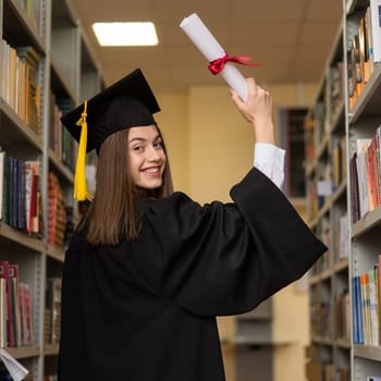 Happy young woman in graduate gown holding diploma in the library