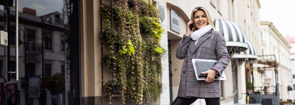 Business woman with a laptop in her hands speaks on a mobile phone in front of the entrance to the office.