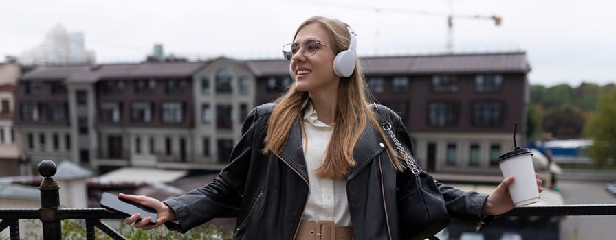 young cheerful woman listening to music in headphones with a cup of coffee in her hands in the city.