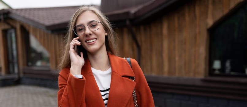 stylish young businesswoman speaks on a mobile phone in a bright orange coat on the background of a modern building.