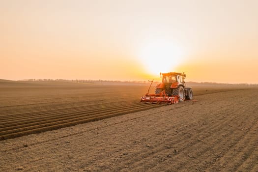 PRAGUE , CZECH REPUBLIC - MARCH 18 2022: Modern tractor cultivates soil in field on agricultural farm at bright sunset. Powerful machine works dragging plow behind at rural site in evening