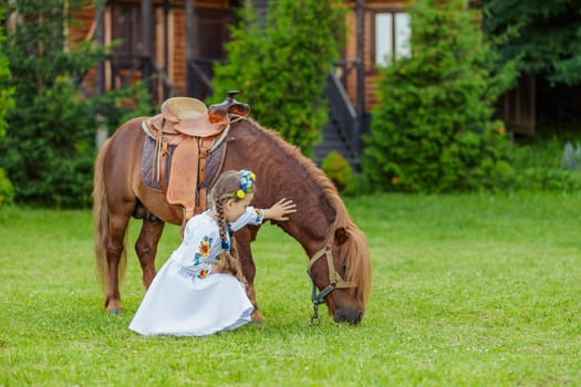 young girl in Ukrainian national dress strokes a pony that grazes on the lawn