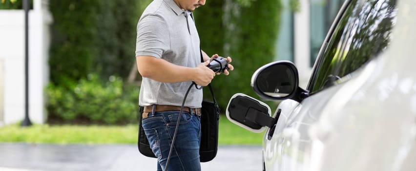 Progressive asian man install cable plug to his electric car with home charging station in the backyard. Concept use of electric vehicles in a progressive lifestyle contributes to clean environment.