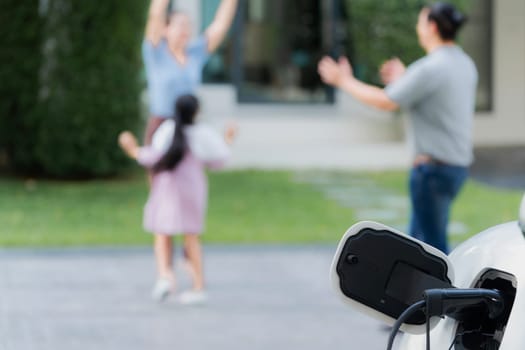 Focus closeup electric vehicle recharging battery from home electric charging station with blurred family in background. Renewable clean energy car for progressive eco awareness lifestyle concept.