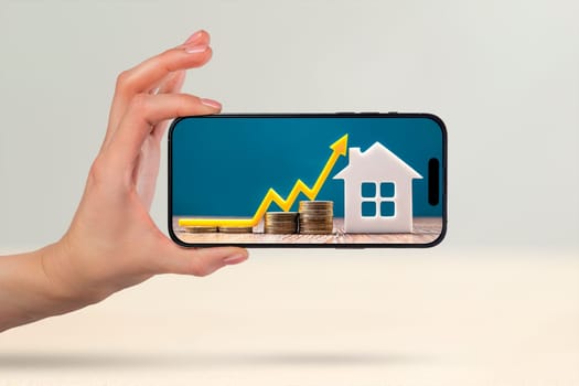 Real estate market, graph, up arrow. House model and stack of coins. Hand with phone and copy space. On the screen of the phone the concept of cost, insurance, real estate mortgage