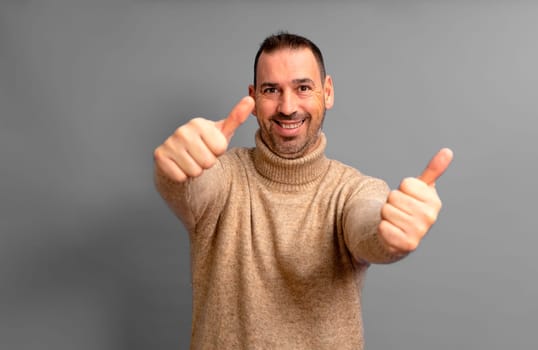 Handsome bearded Hispanic man in his 40s wearing a beige turtleneck with his arms outstretched giving a thumbs up, isolated over gray background